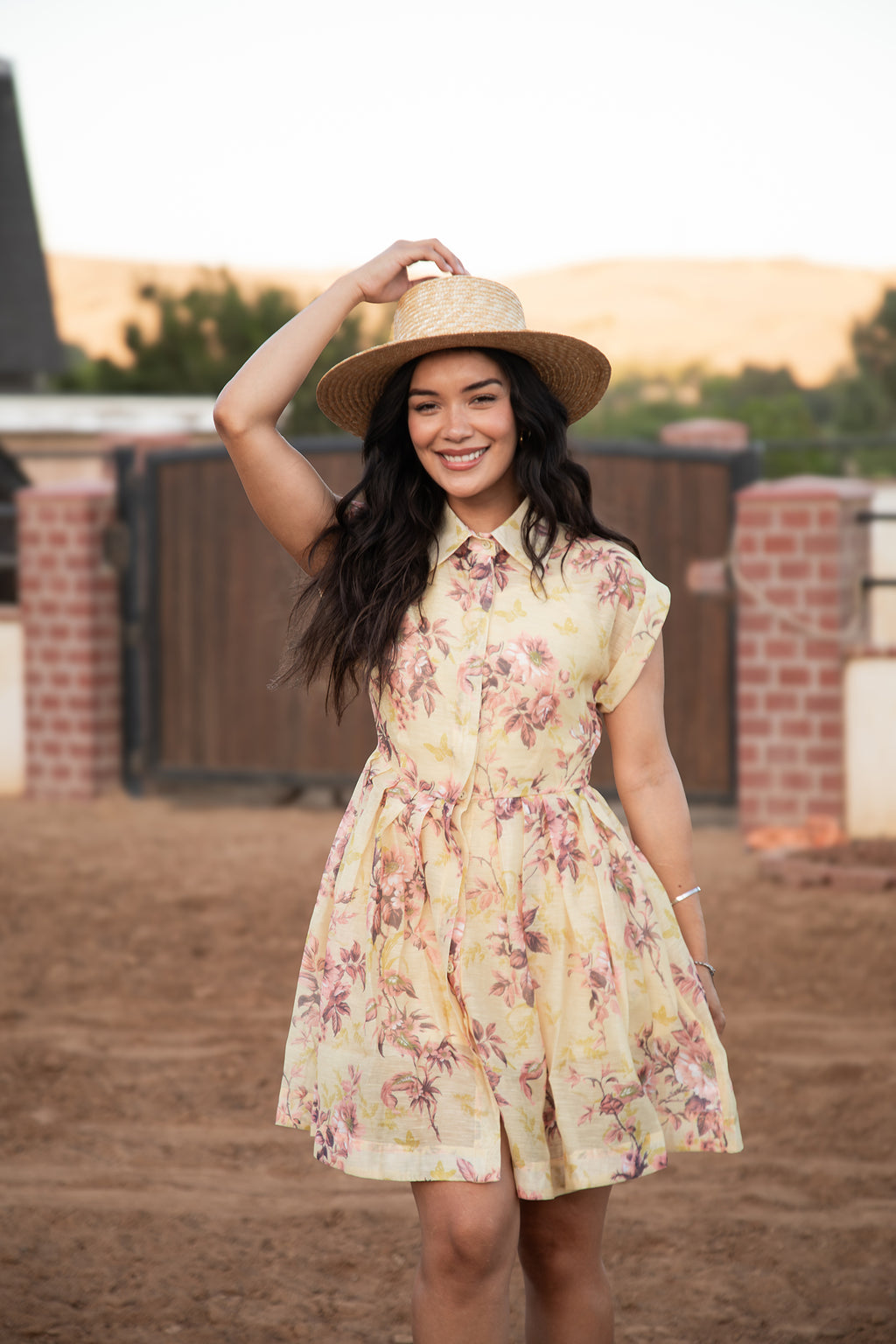Model wearing the Chloe Dress, a buttercream yellow floral shirt dress with a straw hat